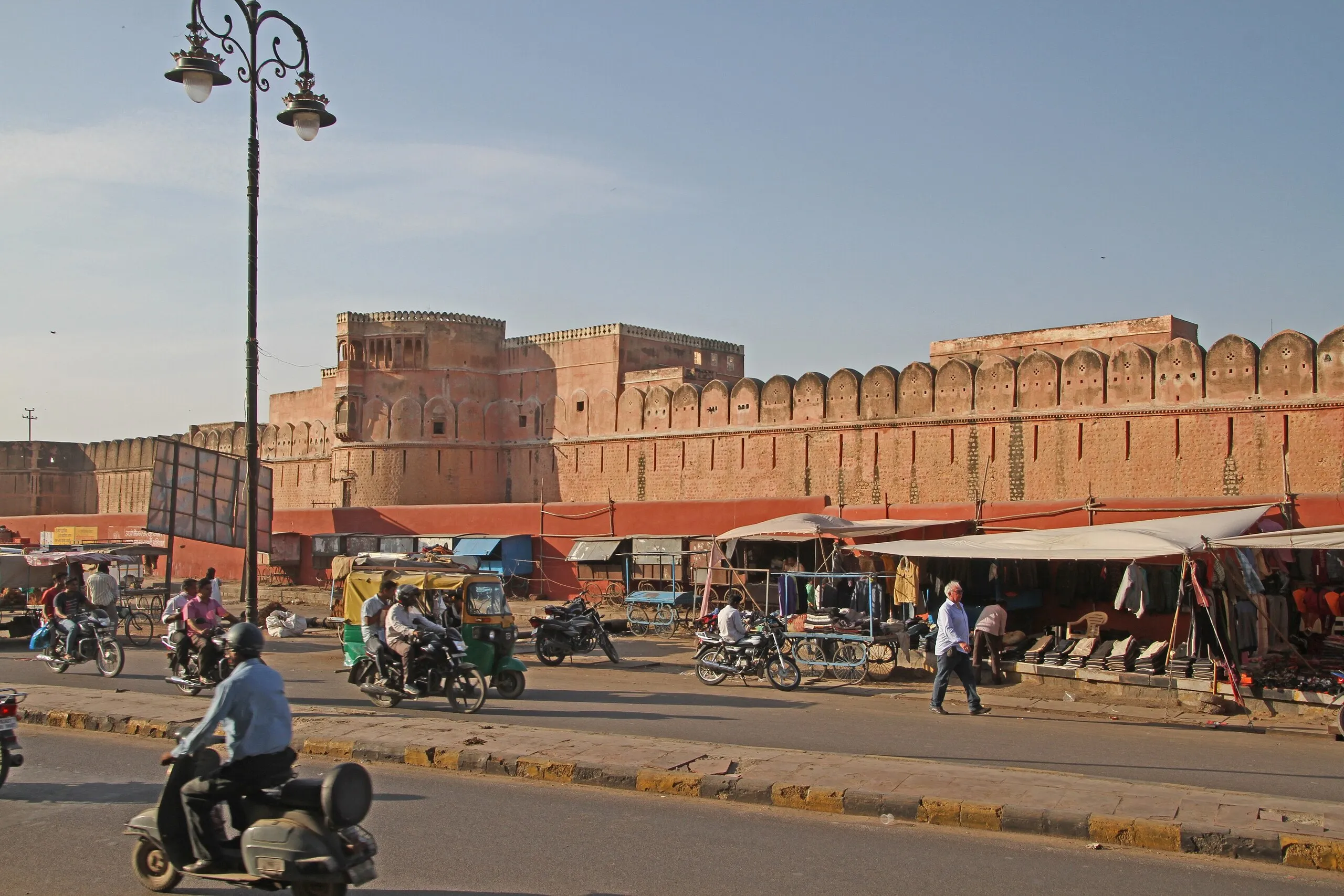 Scooters, an auto-rickshaw and awning clothing stalls on the road below the red sandstone ramparts of Junagarh Fort, Bikaner