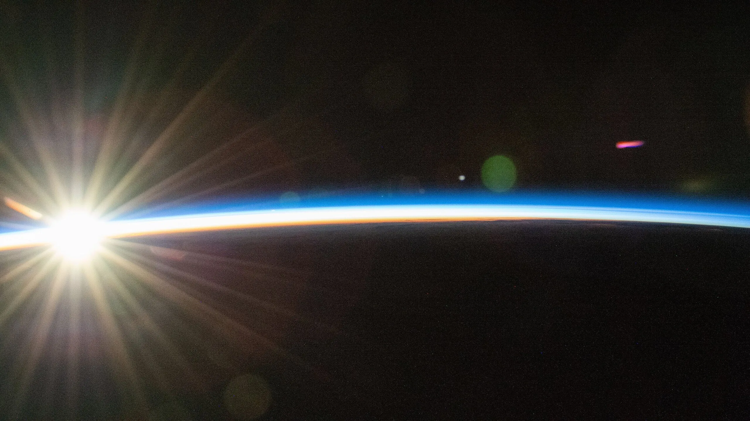 Orbital sunrise from the ISS: a starburst sun at the left edge of Earth's thin blue and orange atmosphere above the Pacific, on black