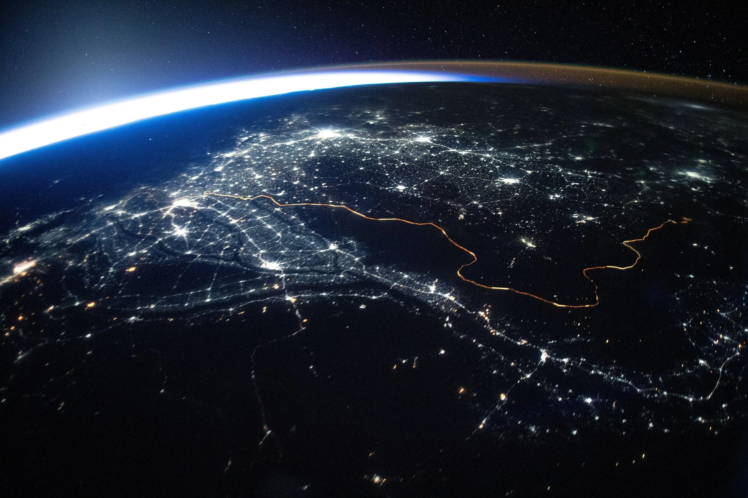 Night view from the ISS of city lights across Pakistan and India, the lit border glowing orange beneath Earth's bright limb