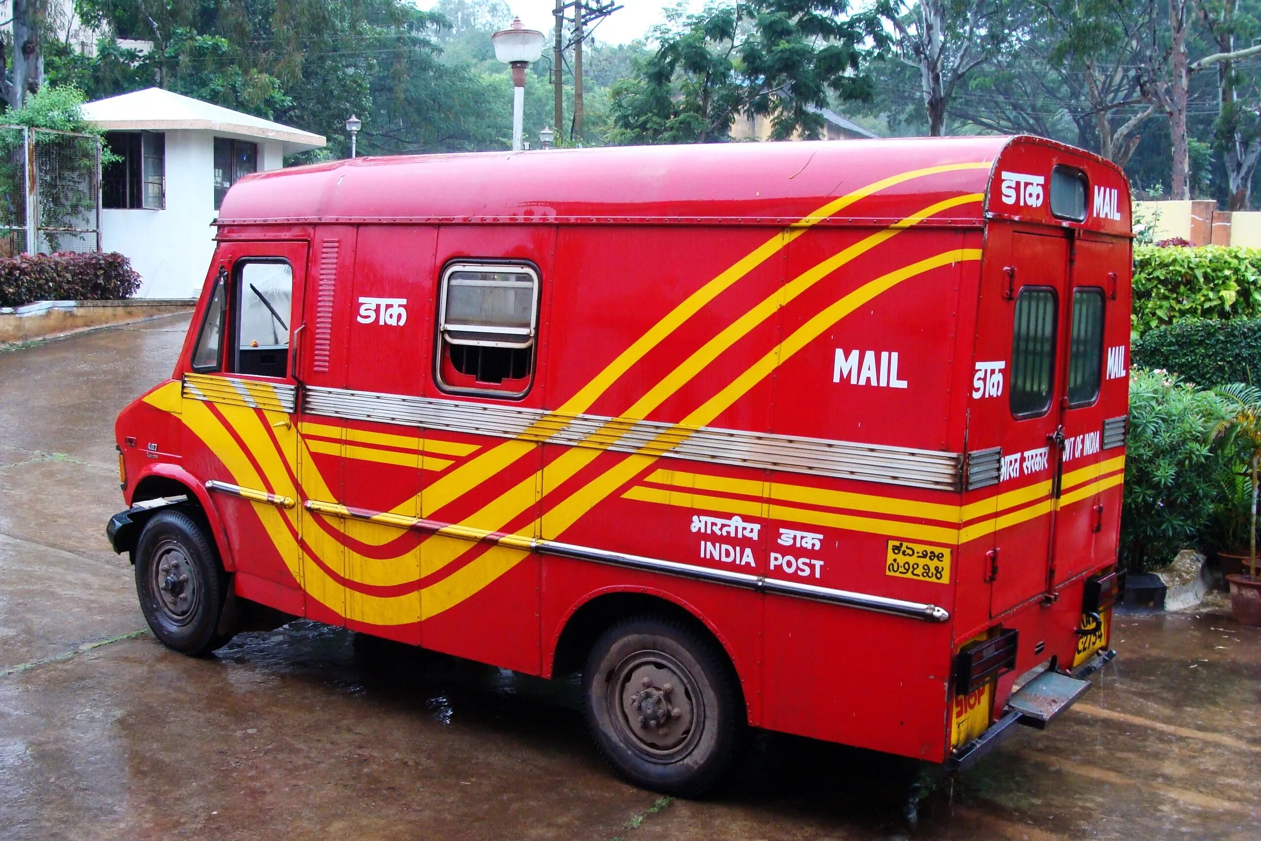 Red-and-yellow India Post mail van parked in the rain in Dharwad, Karnataka, with Hindi and English lettering