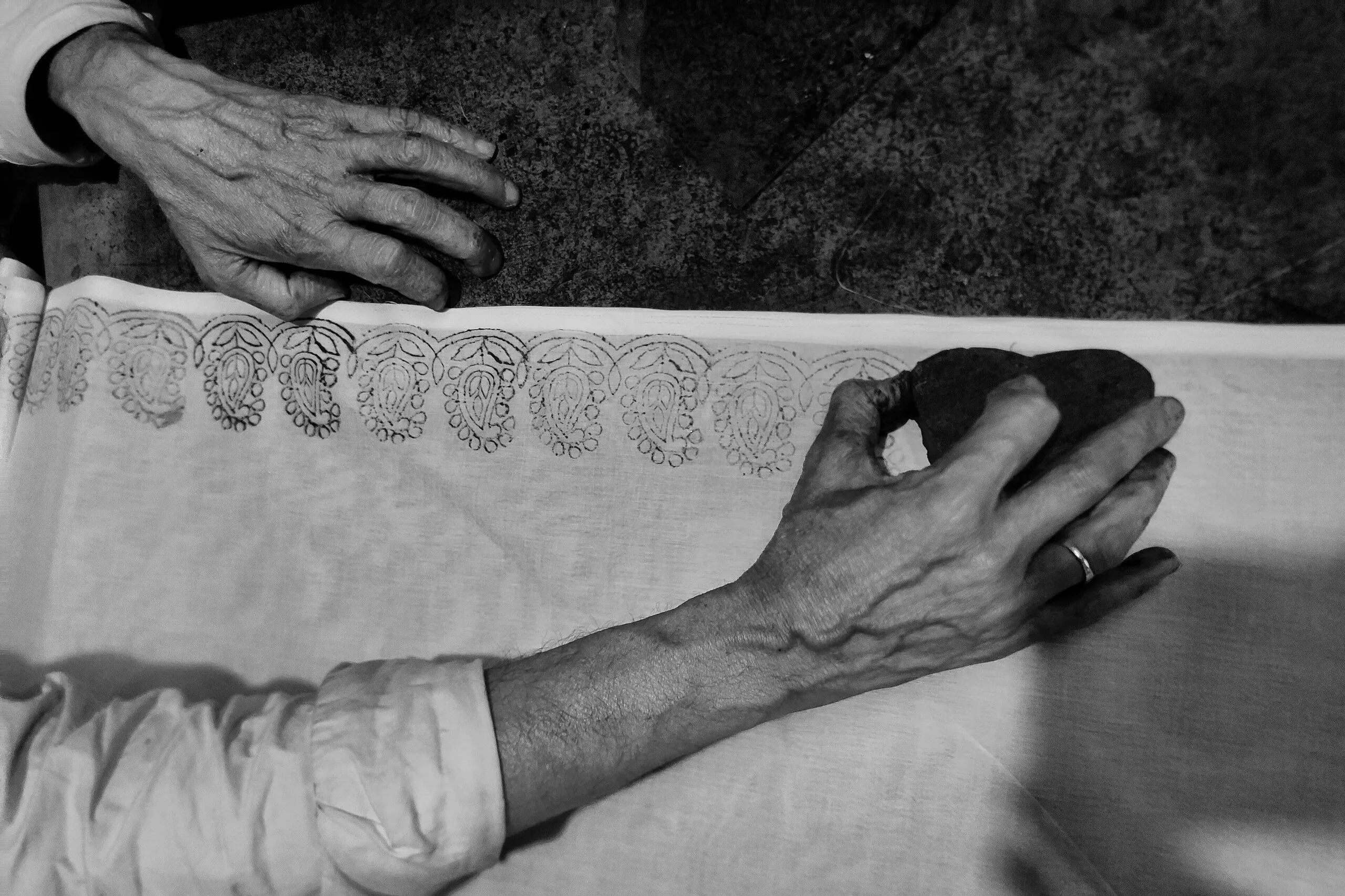 Black-and-white overhead view of an artisan's hands pressing a carved block to print a paisley border onto cotton cloth