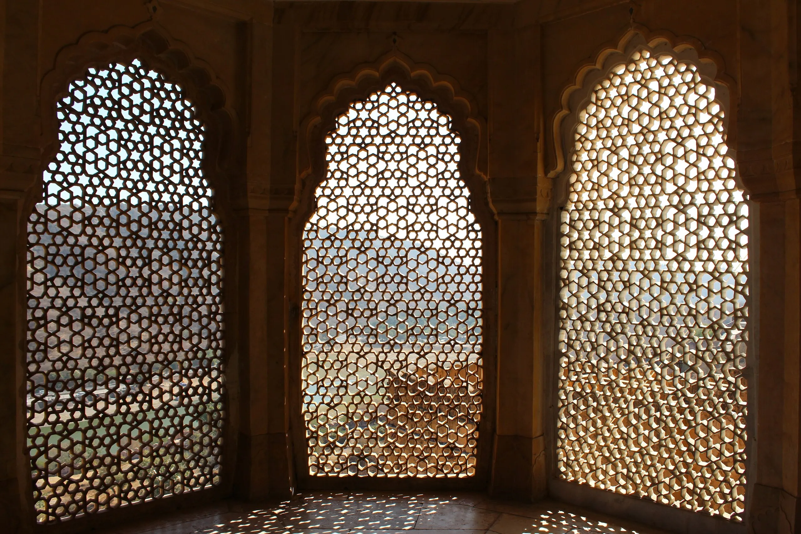 Three arched windows of fine star-pattern stone jaali at Amber Fort, Jaipur, sunlight dappling the floor and hills beyond