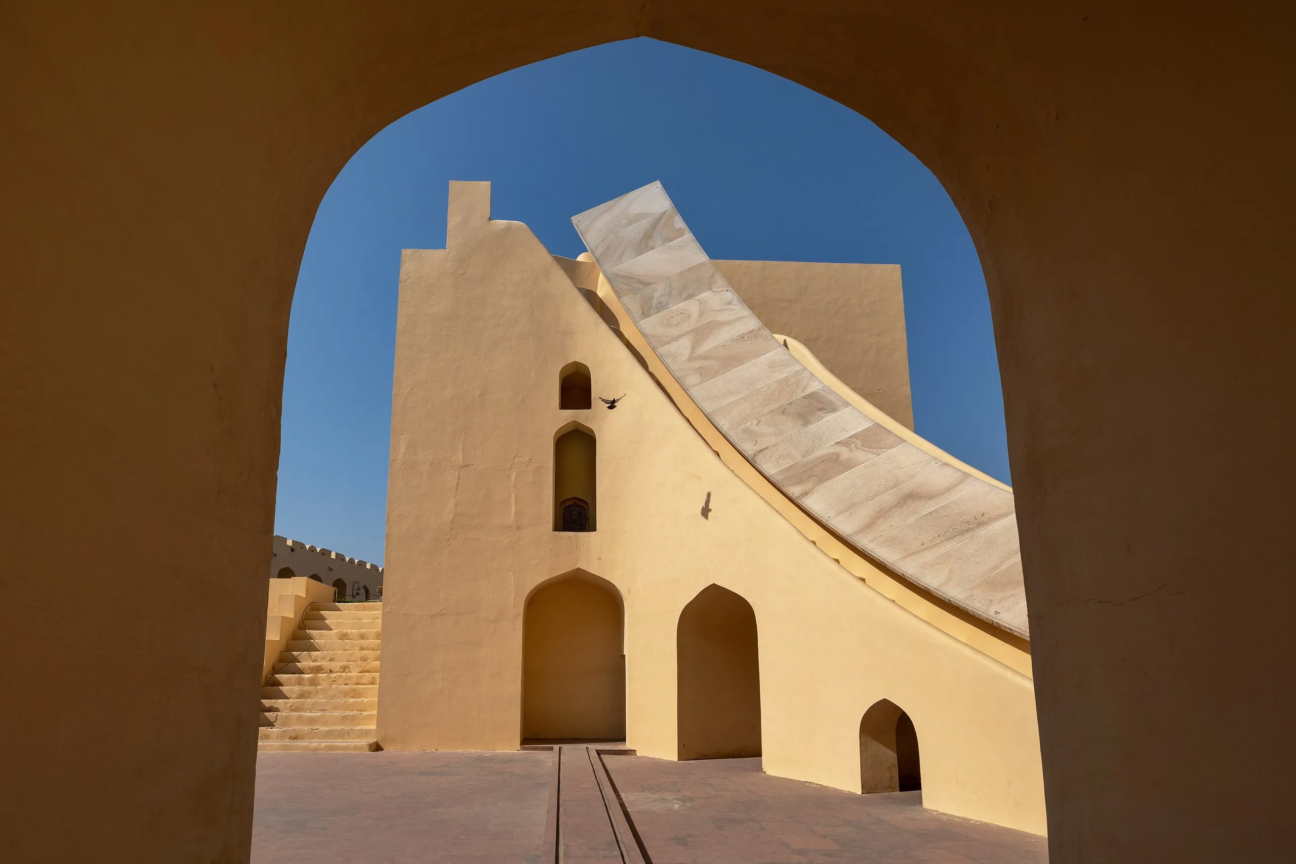 A yellow-ochre Jantar Mantar instrument with its curved stone stair, framed by a dark arched doorway against a deep blue sky