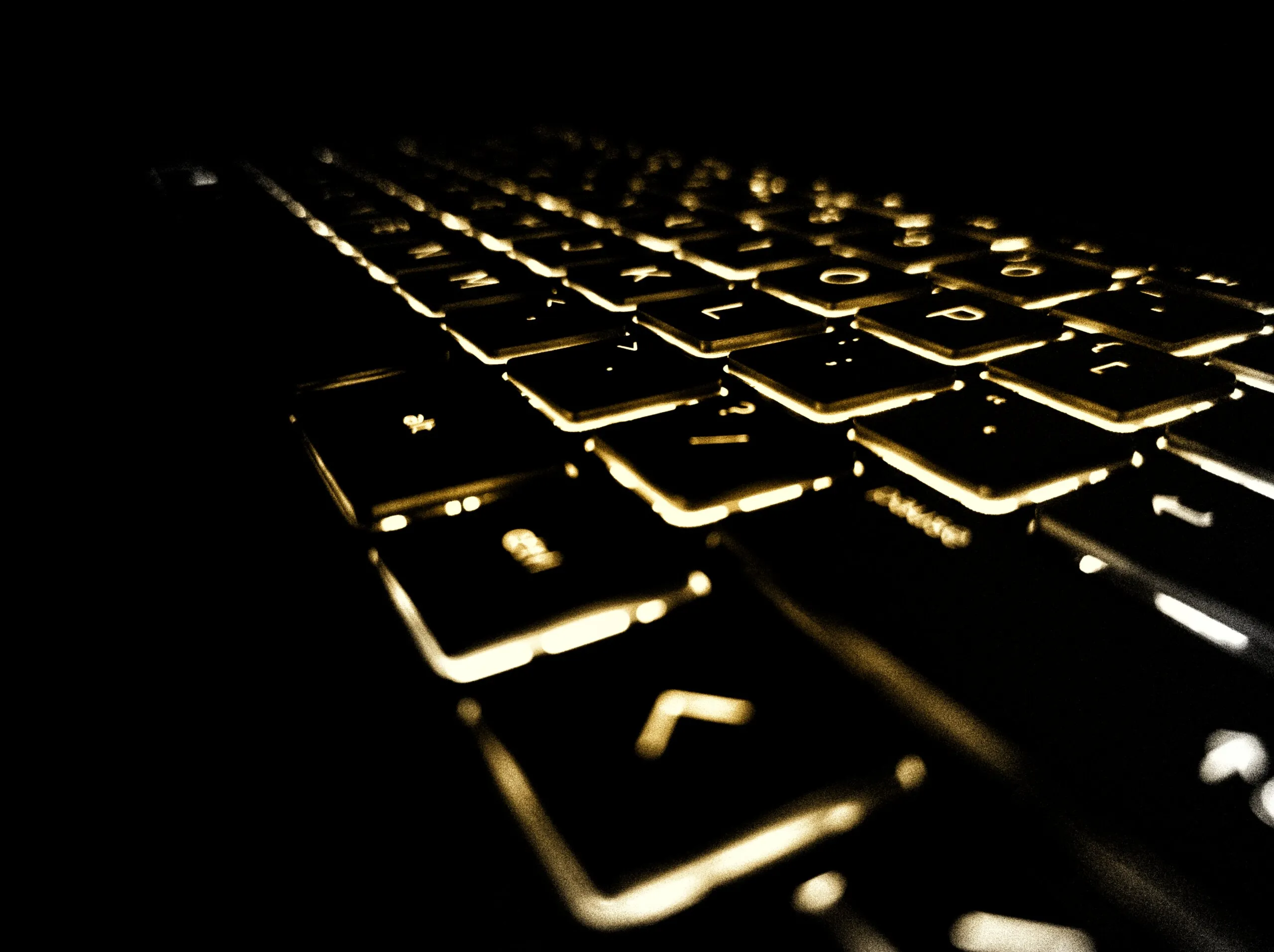 Backlit laptop keyboard shot at a low angle, key edges and legends glowing gold against pure black