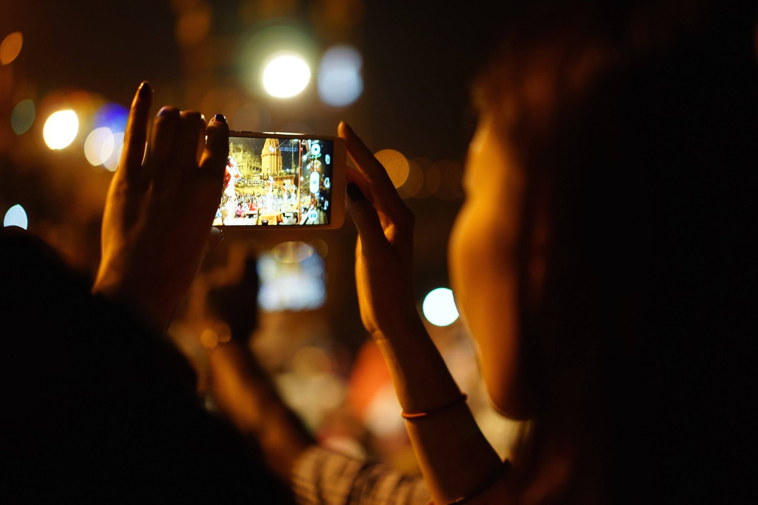 Hands holding up a smartphone to film the evening Ganga aarti at Dasashwamedh Ghat, Varanasi, against warm bokeh lights