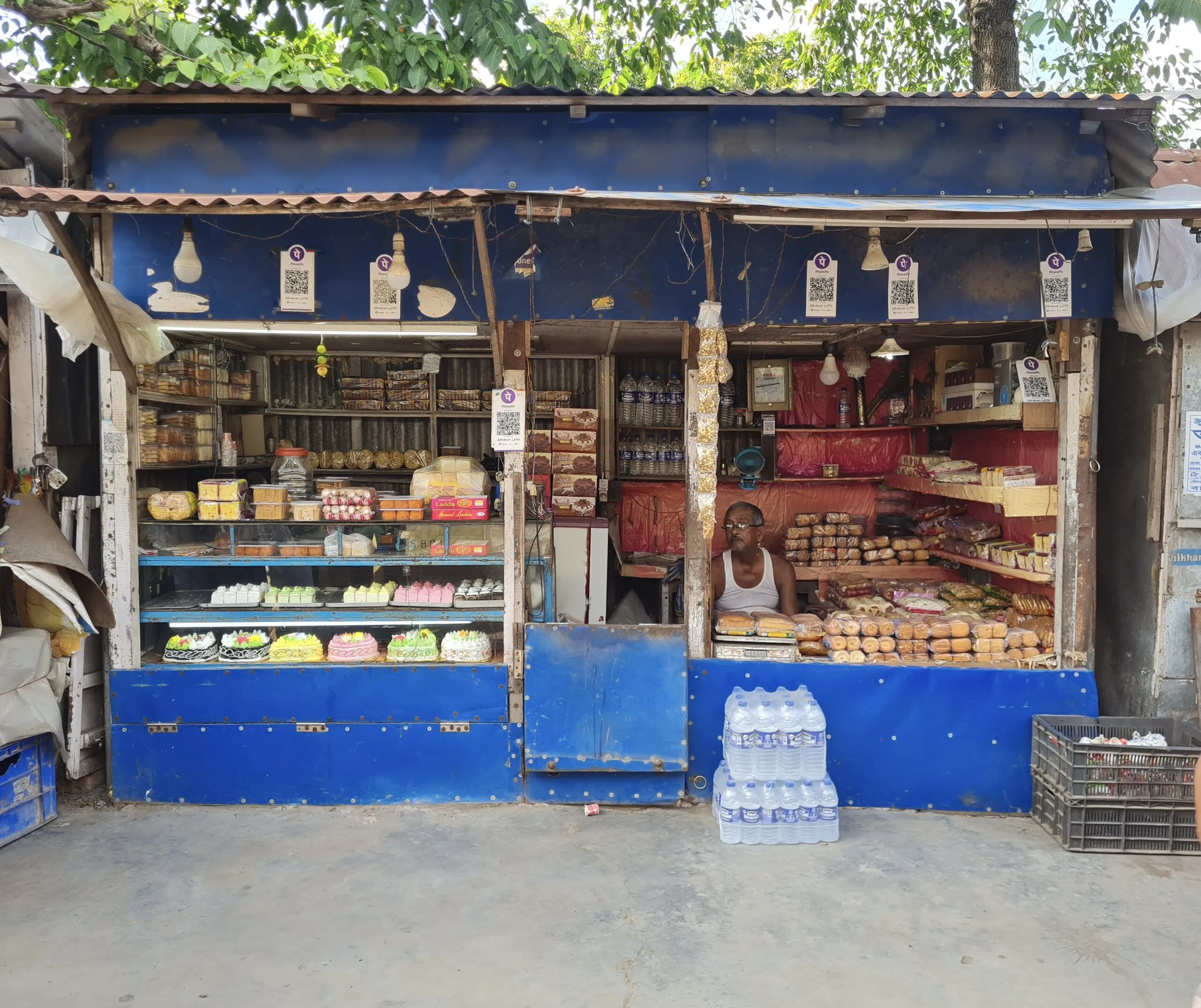Blue tin-roofed sweet and bakery stall in North 24 Parganas, West Bengal, with four PhonePe UPI QR placards above the counter
